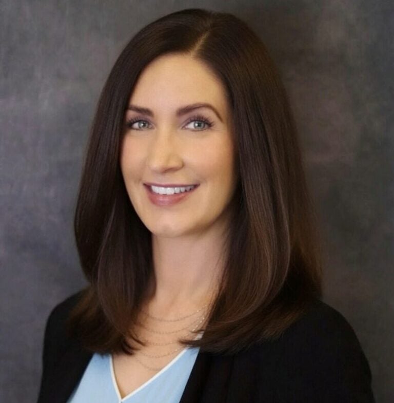 Confident professional woman with long brown hair smiling, wearing a black blazer and light blue top, against a neutral background.